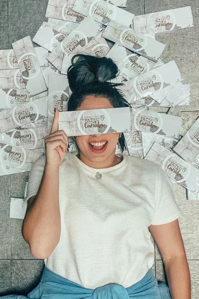 Pessoa deitada no chão cercada por dezenas de vouchers do Tour O Que Fazer Curitiba, segurando um deles sobre os olhos.